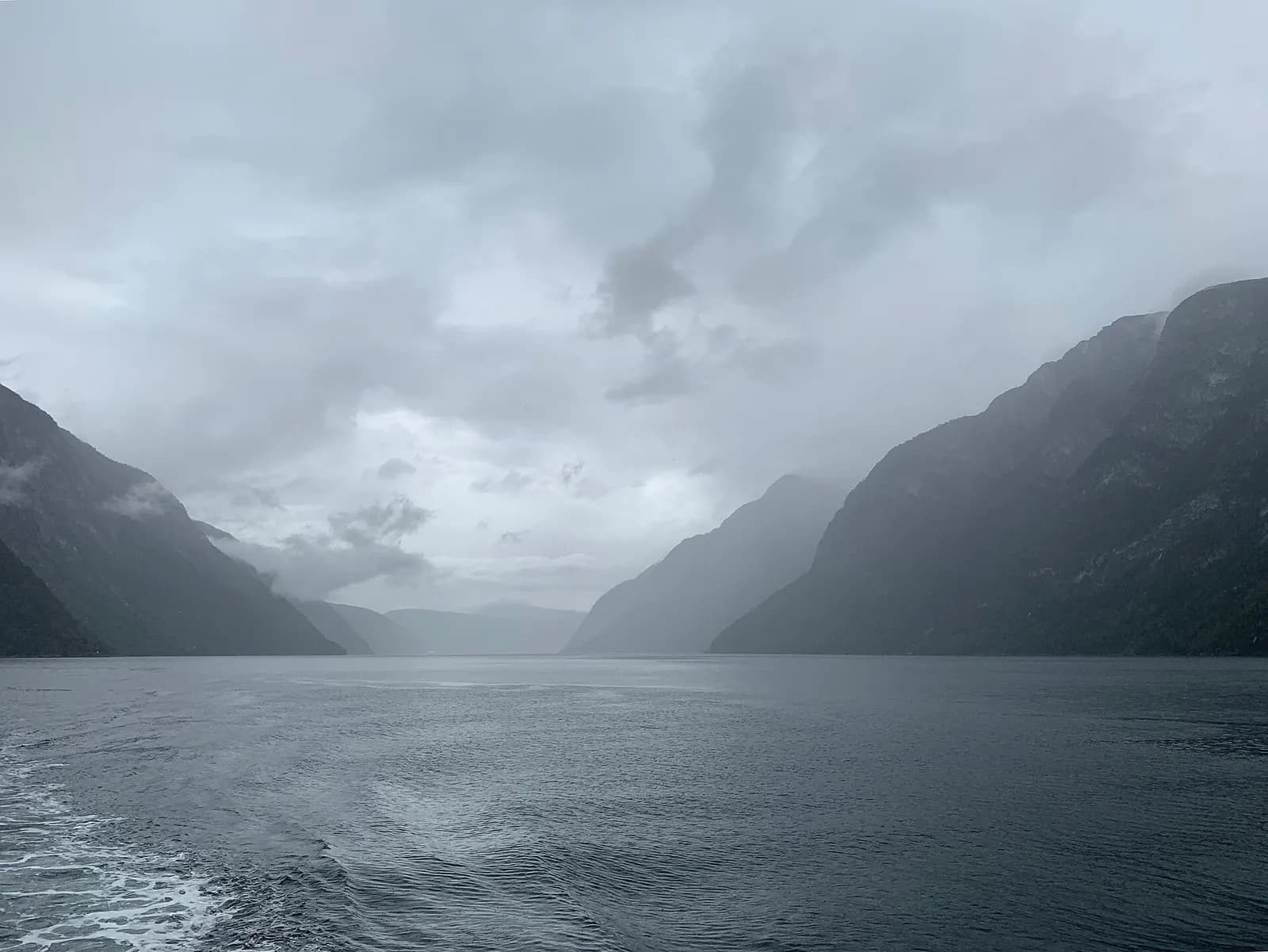 vue depuis l'arrière d'un bateau sur le fjord, entre des montagnes sous un ciel gris