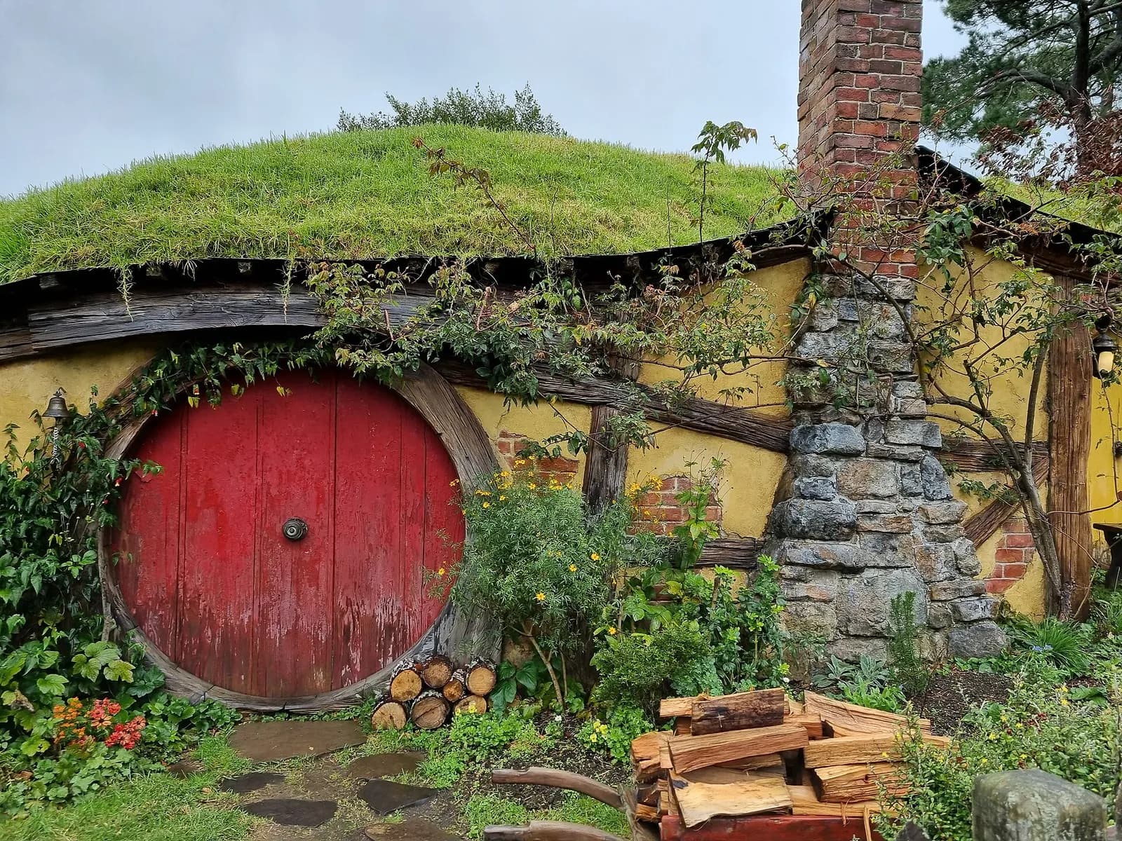 une porte ronde rouge et une cheminée en pierre sur une maison de hobbit fleurie