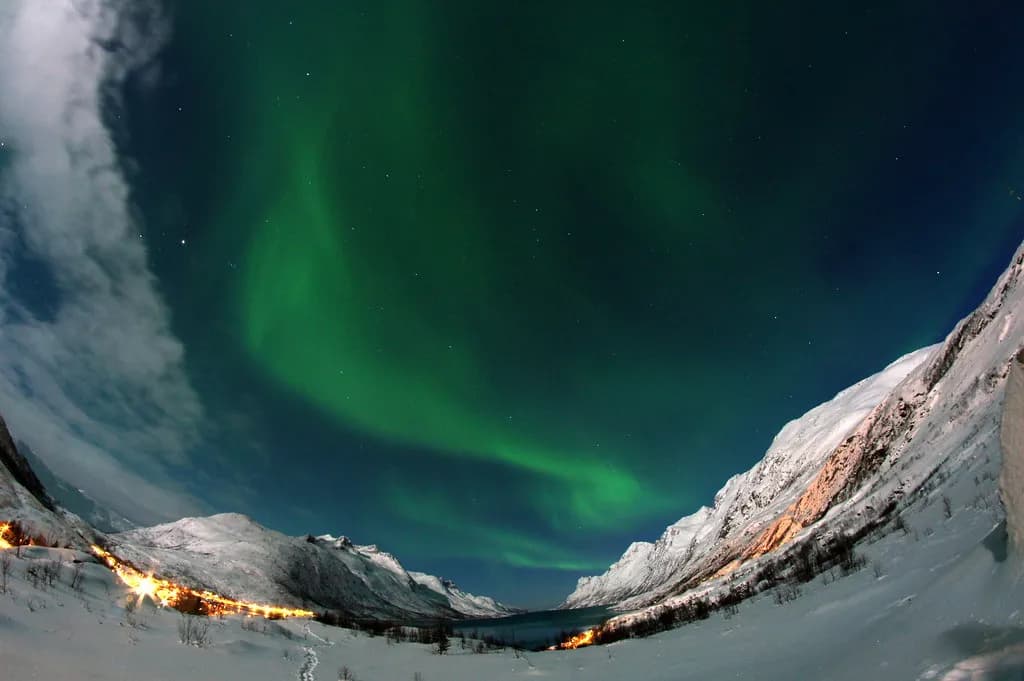 aurore boréale au-dessus d'un fjord bordé de montagnes enneigées et de maisons éclairées