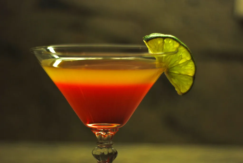 an orange and red layered cocktail in a martini glass with lime