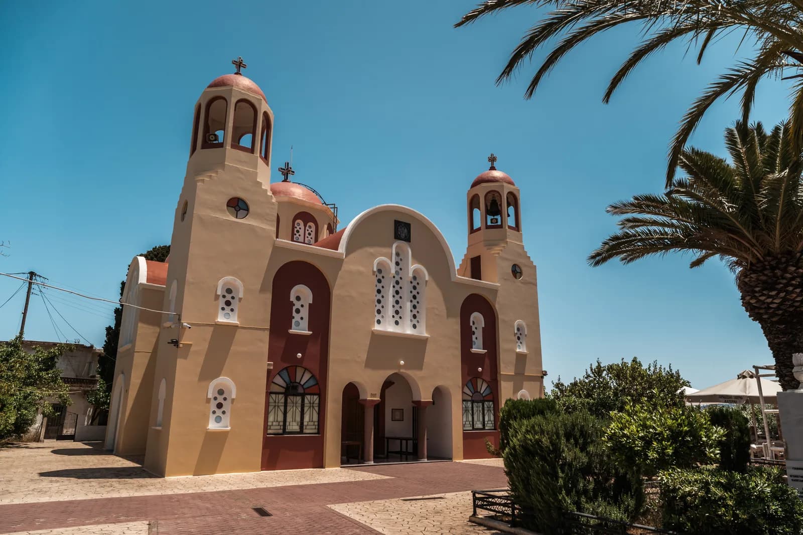 Église orthodoxe aux deux clochers et aux façades ocre et bordeaux, entourée de palmiers, dans un village crétois