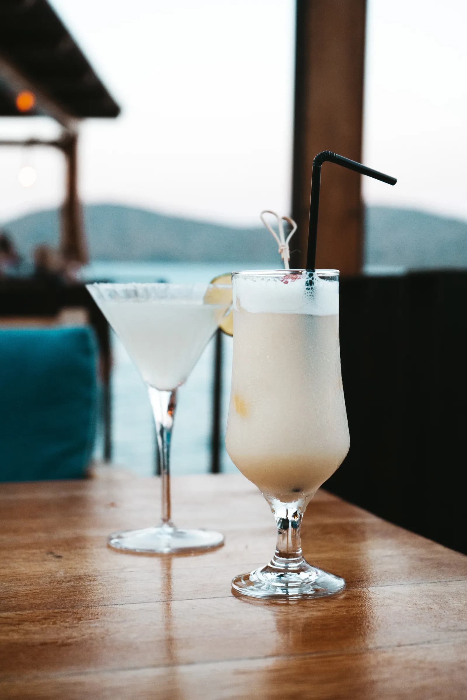 Two cocktails on a wooden taverna table, with the bay of Elounda in the background