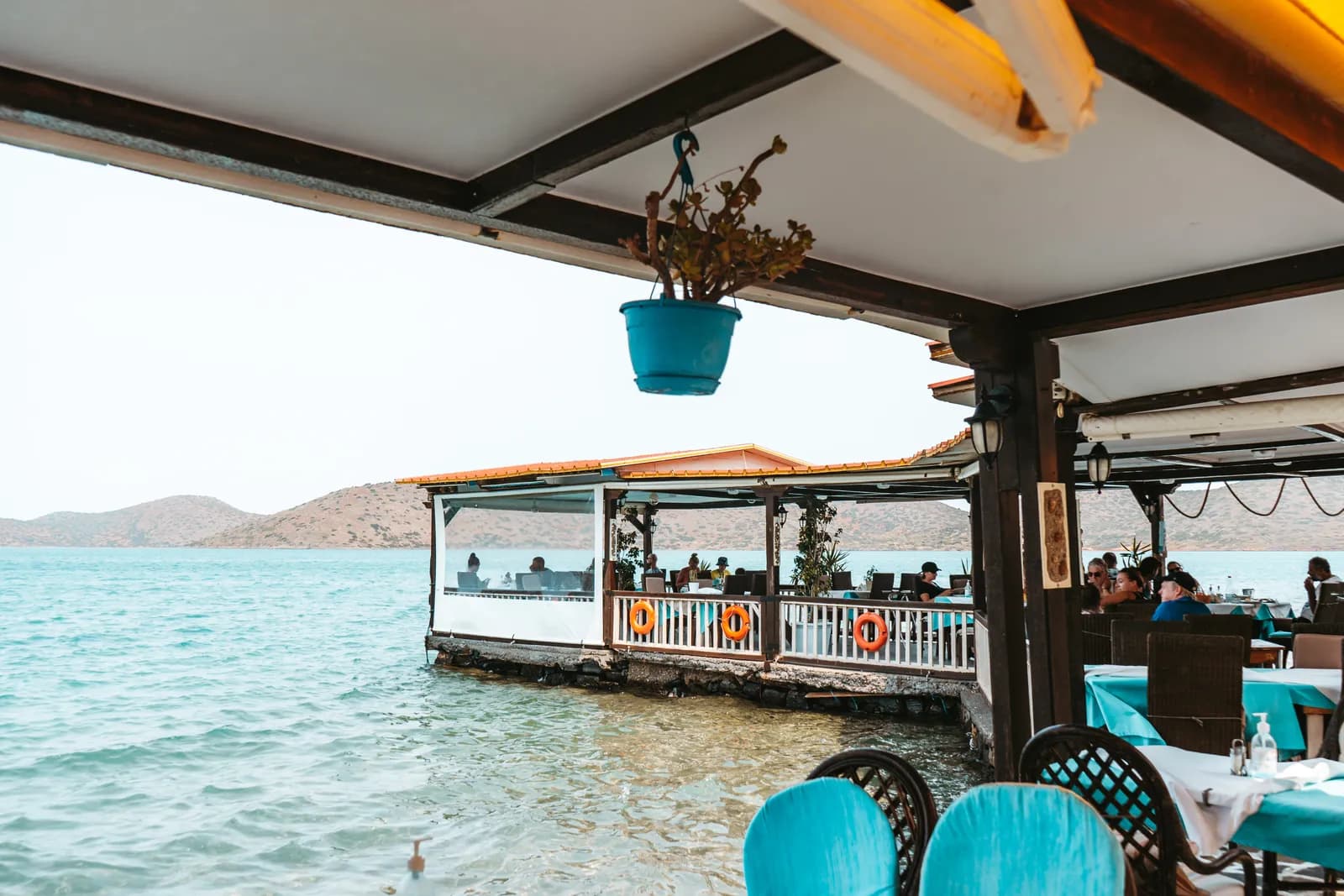 Terrasse de taverne posée sur l'eau à Elounda, chaises turquoise et salle sur pilotis face au golfe