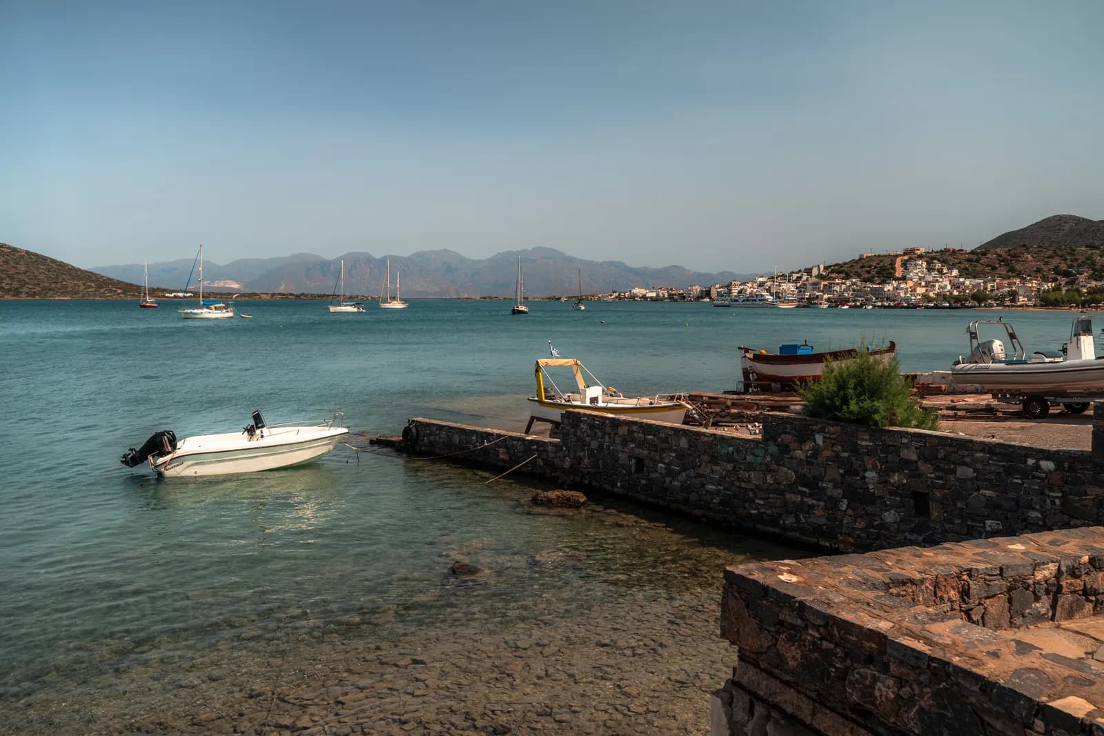 Barques et jetée de pierre sur le rivage, avec le village d'Elounda de l'autre côté de la baie