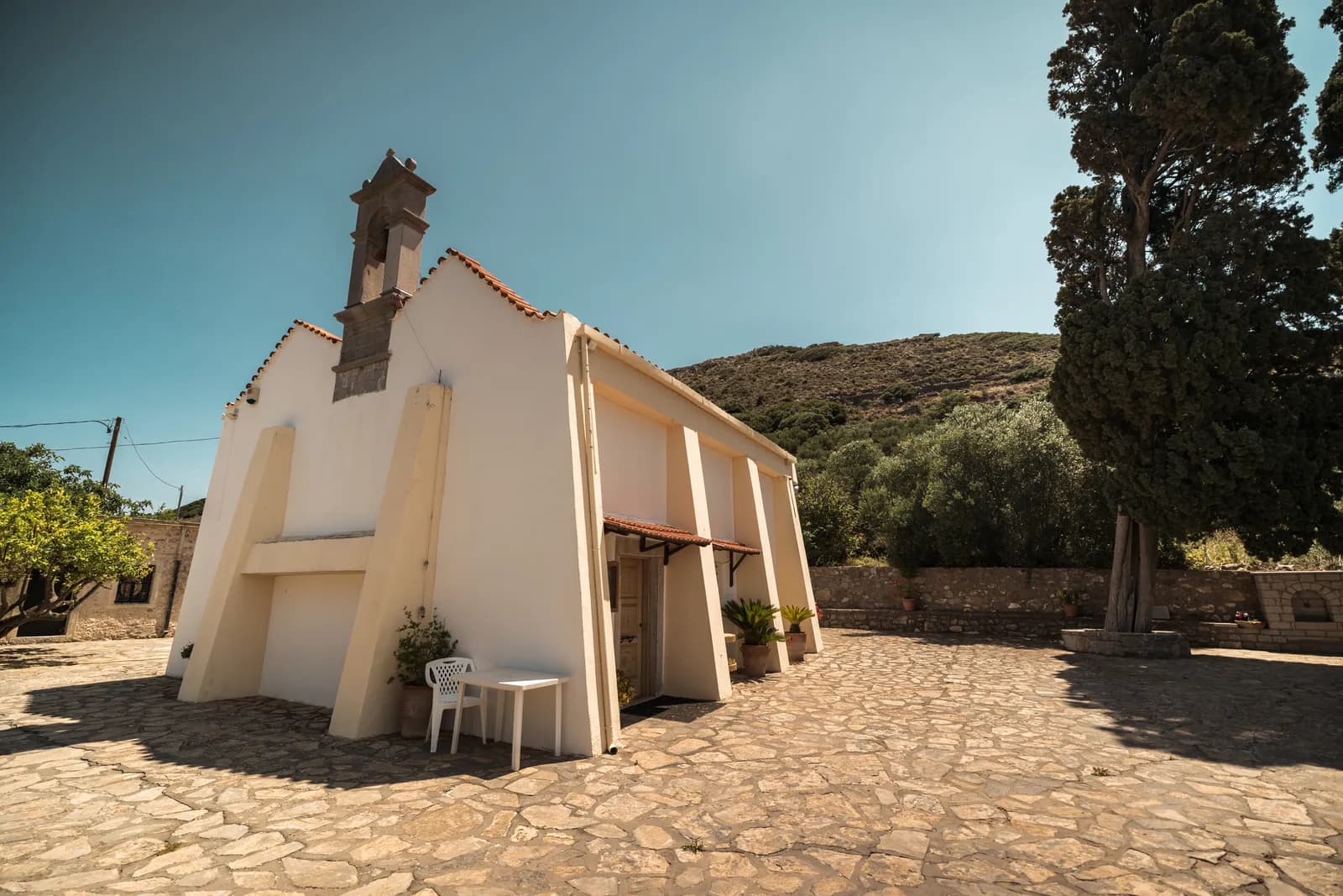 Petite église blanche et grand cyprès sur une place dallée de village, dans l'arrière-pays d'Elounda