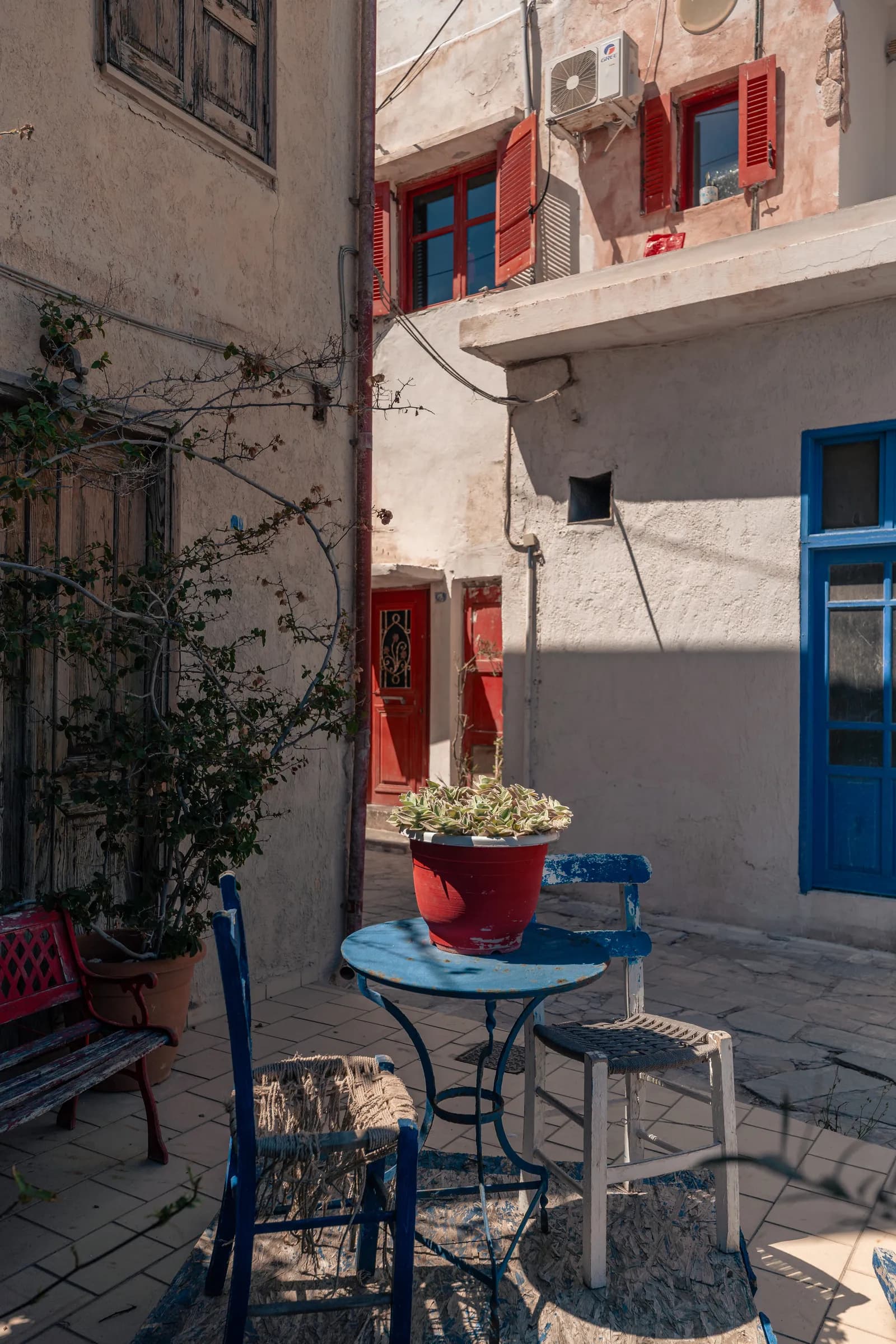 Petite table bleue, chaises paillées et volets rouges dans une cour du vieux quartier de Kato Mera à Ierapetra
