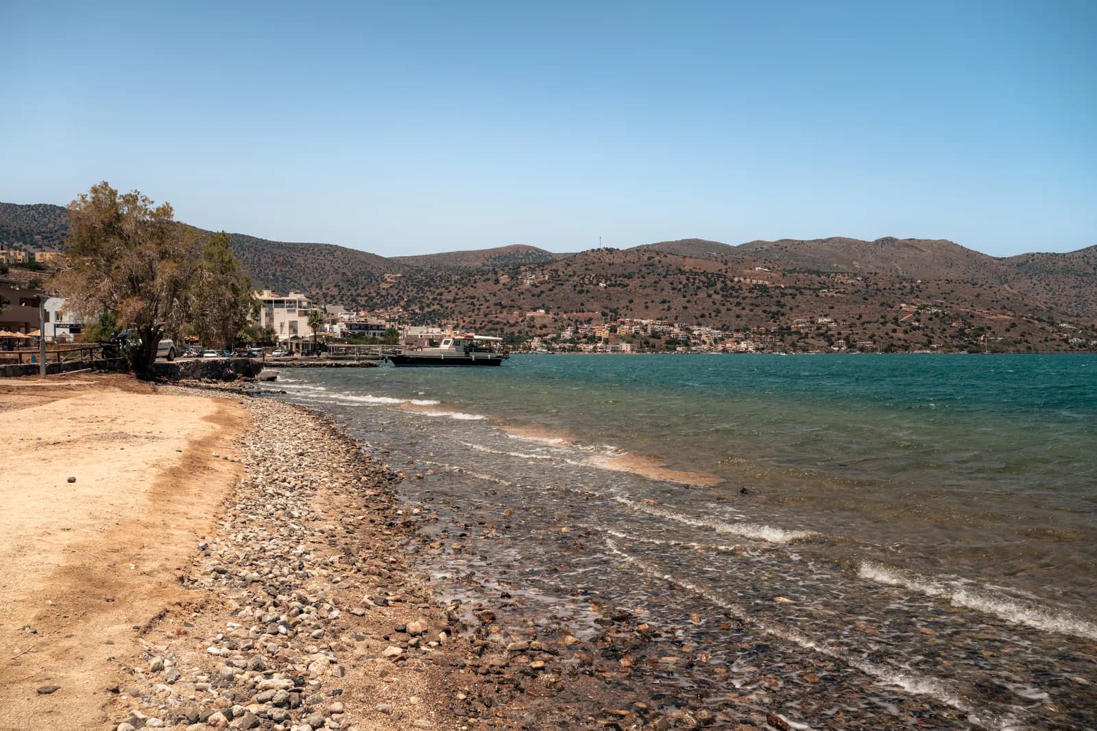 La plage de sable et de galets d'Elounda, avec le port et les collines de la presqu'île au loin
