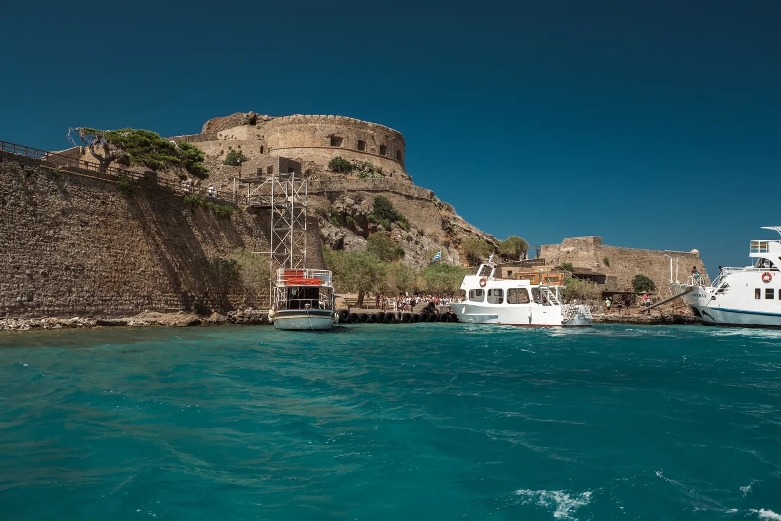 Bateaux de passeurs accostés au pied des remparts vénitiens de Spinalonga, dans une eau turquoise