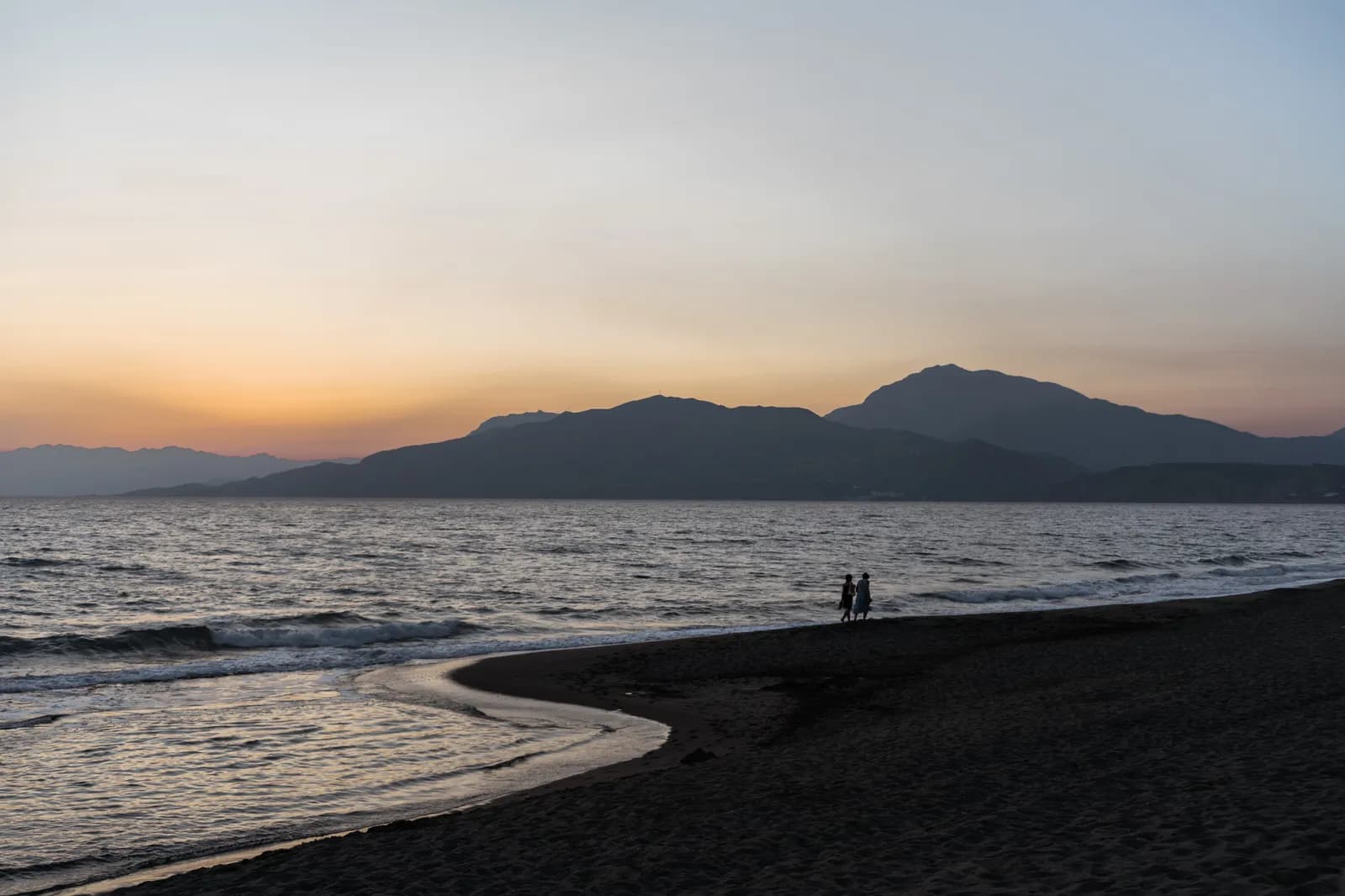 Plage de sable sombre de la côte sud au crépuscule, promeneurs lointains et montagnes en silhouette