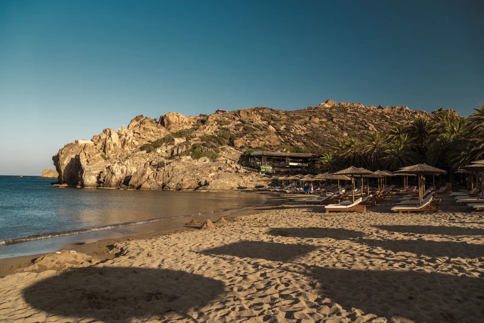 La plage de sable de Vai en fin de journée, ses transats sous les paillotes et son rocher doré