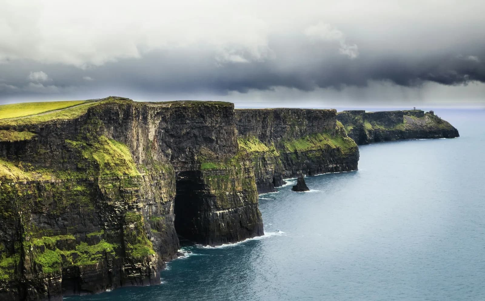 Falaises de Moher sous un ciel d'orage, avec une grotte marine et un piton rocheux au pied des parois