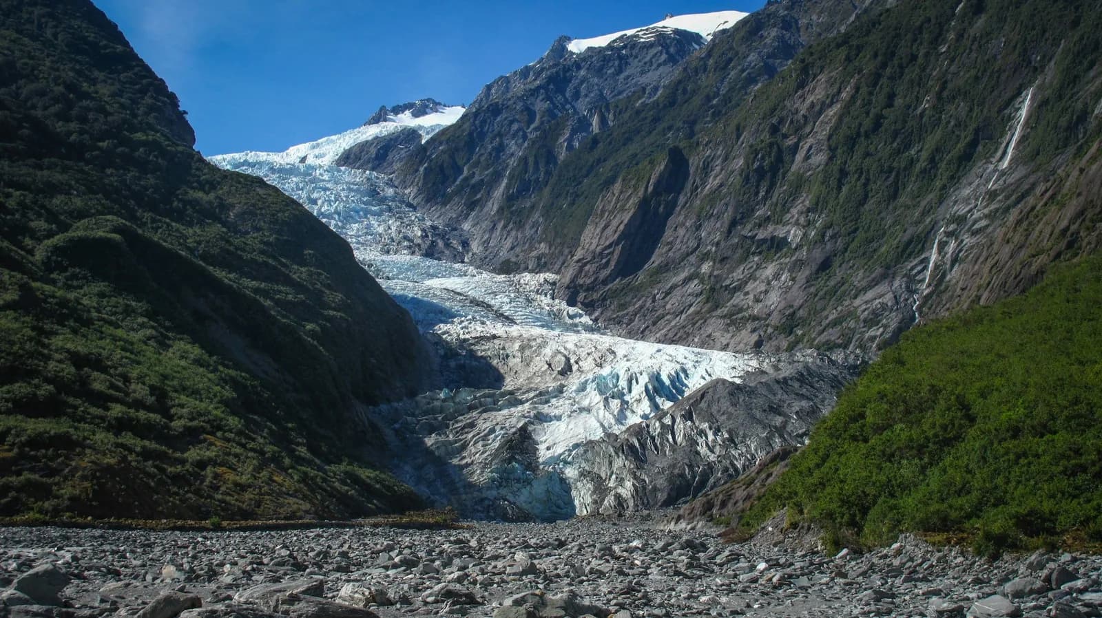 Franz Josef Glacier descending between rainforest-clad valley walls above a boulder-strewn riverbed