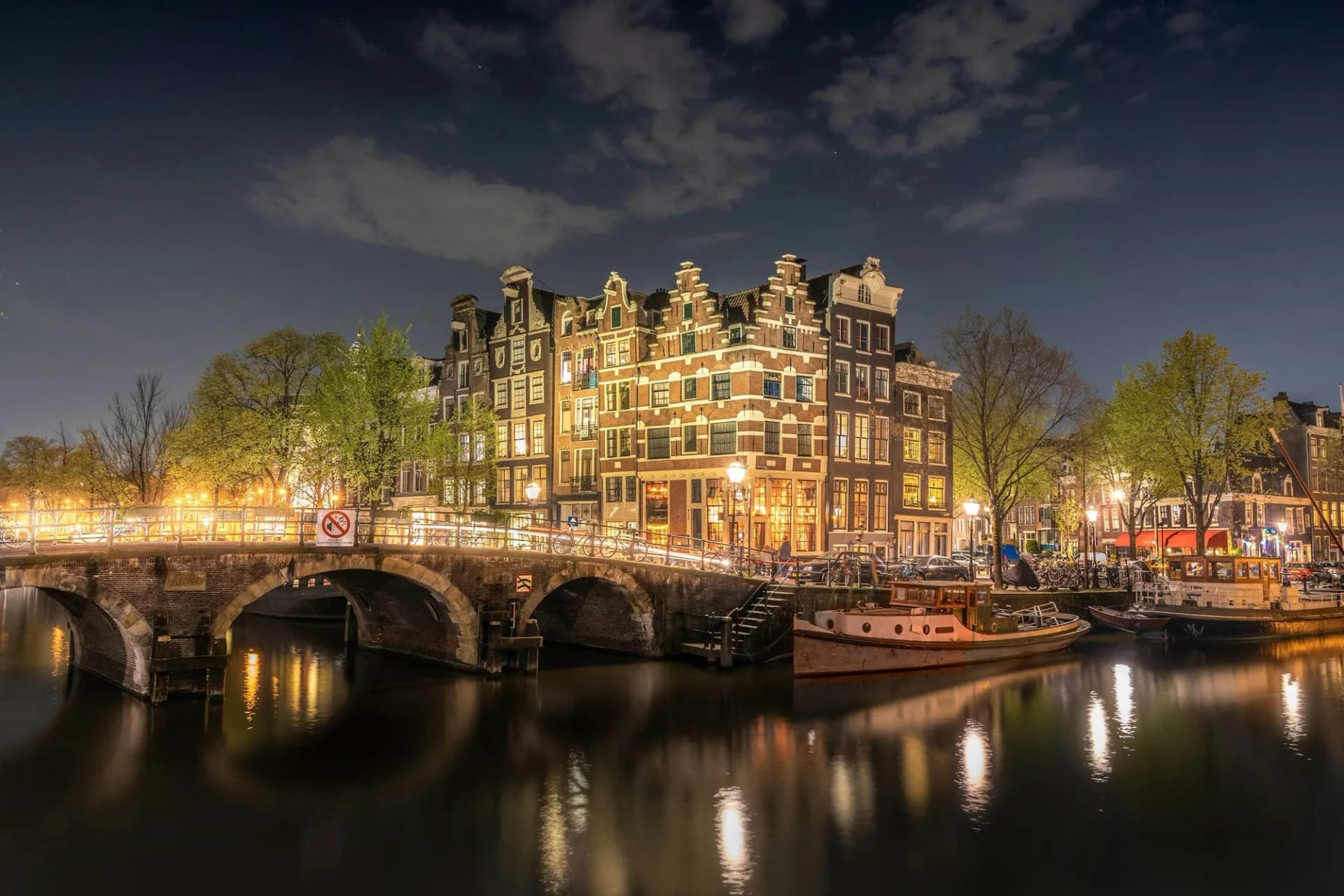 Maisons à pignons illuminées et pont en brique sur un canal d'Amsterdam la nuit, reflets dorés sur l'eau