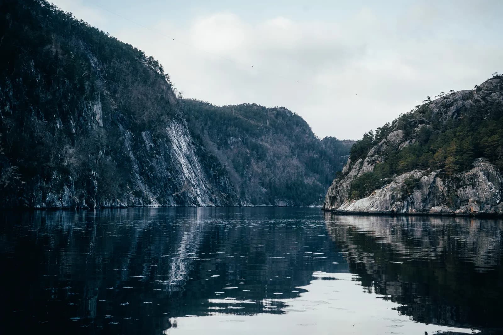 falaises reflétées dans l’eau calme du fjord sous un ciel gris, pendant la croisière vers Mostraumen en mars