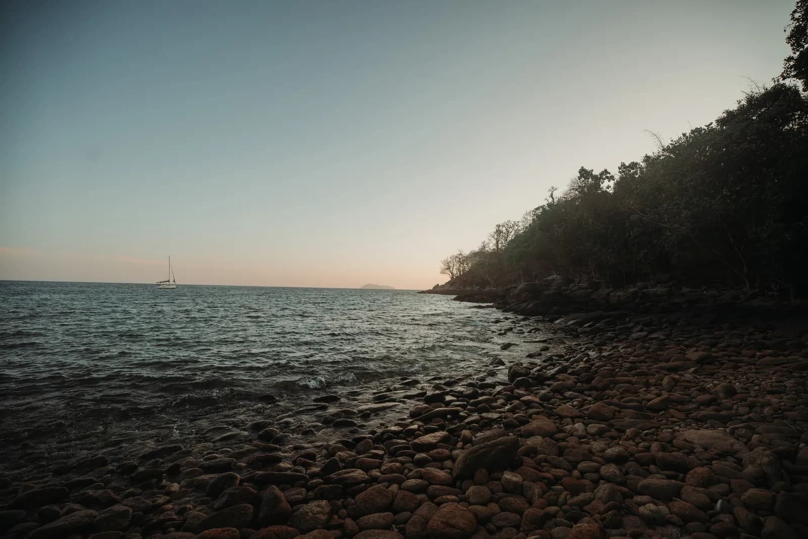Pointe rocheuse et plage de galets de Koh Racha Yai au coucher du soleil, voilier à l'horizon
