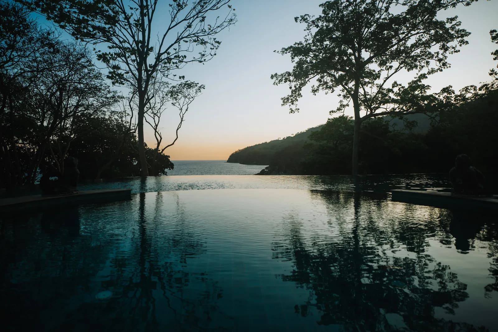 Piscine à débordement d'un resort de Koh Racha au crépuscule, arbres et cap reflétés dans l'eau immobile
