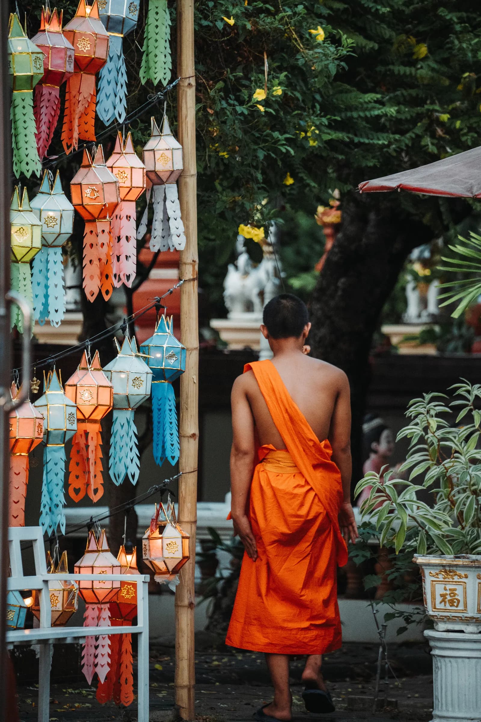 Jeune moine en robe orange vu de dos devant des lanternes lanna, dans un temple de la vieille ville de Chiang Mai