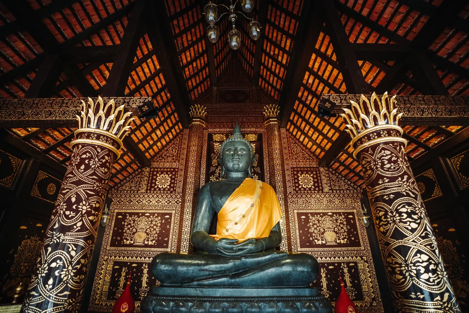 Bouddha de pierre sombre drapé de jaune entre deux colonnes peintes, dans une salle en bois de Wat Chedi Luang